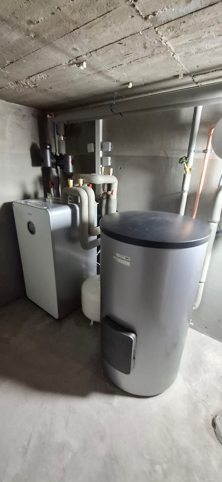Wärmepumpen-Inneneinheit mit Pufferspeicher in grauem Technikraum, isolierte Rohre verlaufen an Wand und Decke.